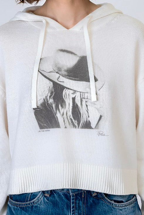 A person wearing a cropped ivory knit hoodie with a large black-and-white "On the Verge Randi" graphic print of a woman in a hat on the front, paired with blue jeans against a plain light background.
