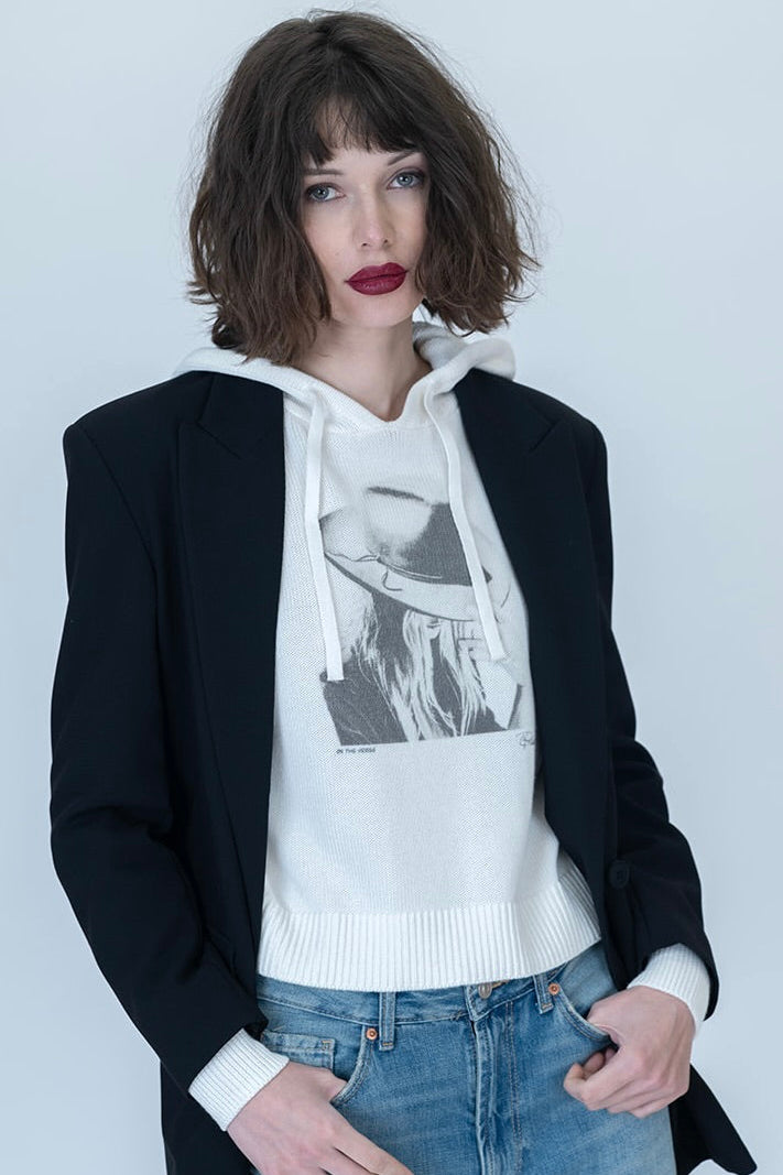 A model wearing a black blazer over a white graphic "On the Verge Randi" hoodie and light-wash distressed blue jeans, standing against a light gray studio background.