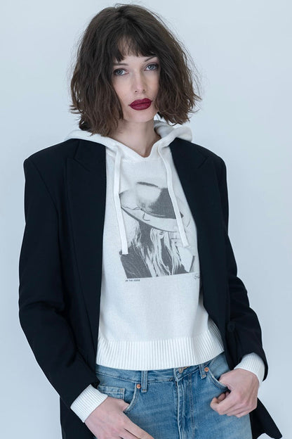 A model wearing a black blazer over a white graphic "On the Verge Randi" hoodie and light-wash distressed blue jeans, standing against a light gray studio background.