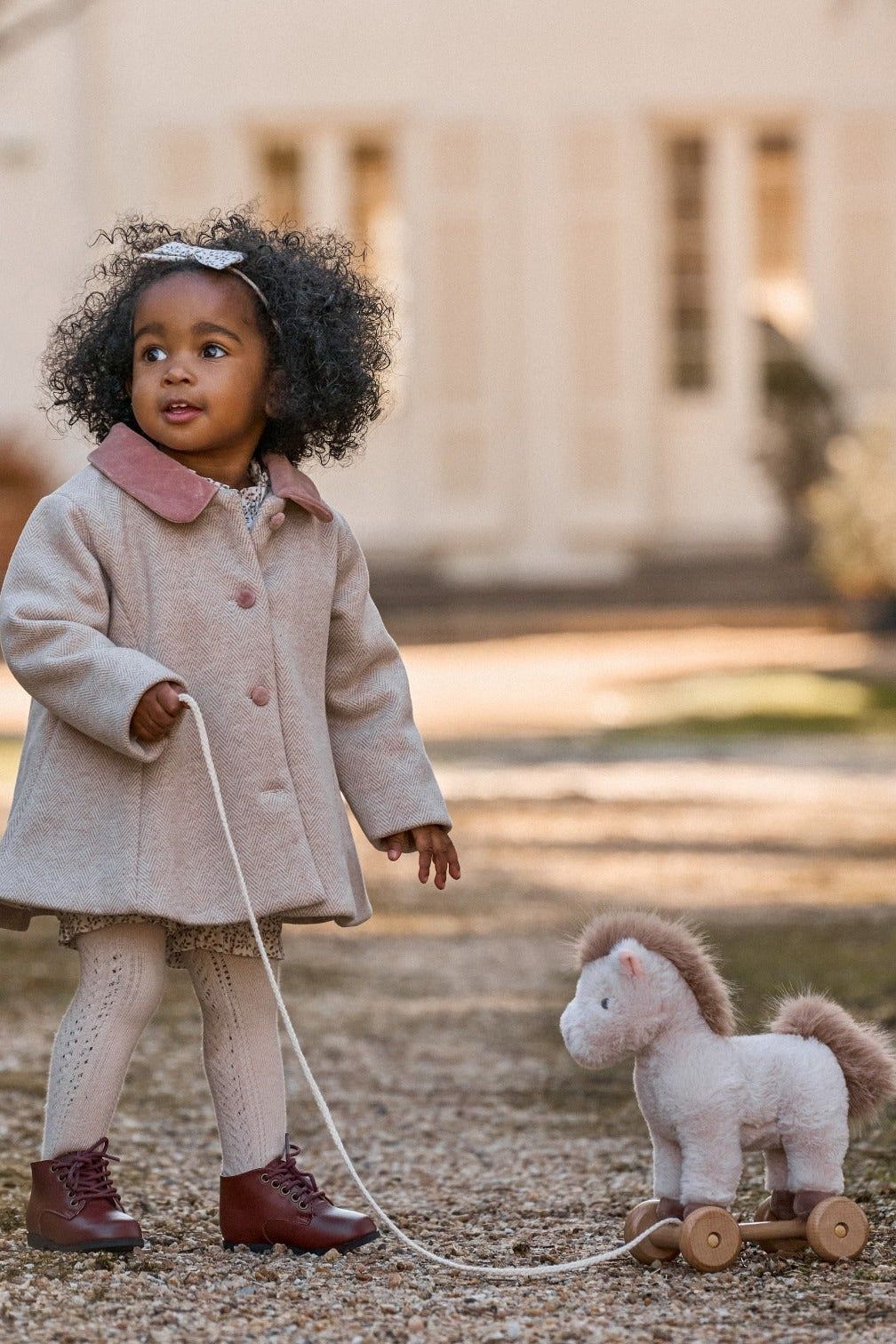 Child in a beige coat pulling a toy horse on a leash outdoors.