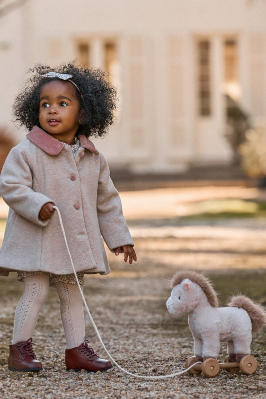 Child in a beige coat pulling a toy horse on a leash outdoors.