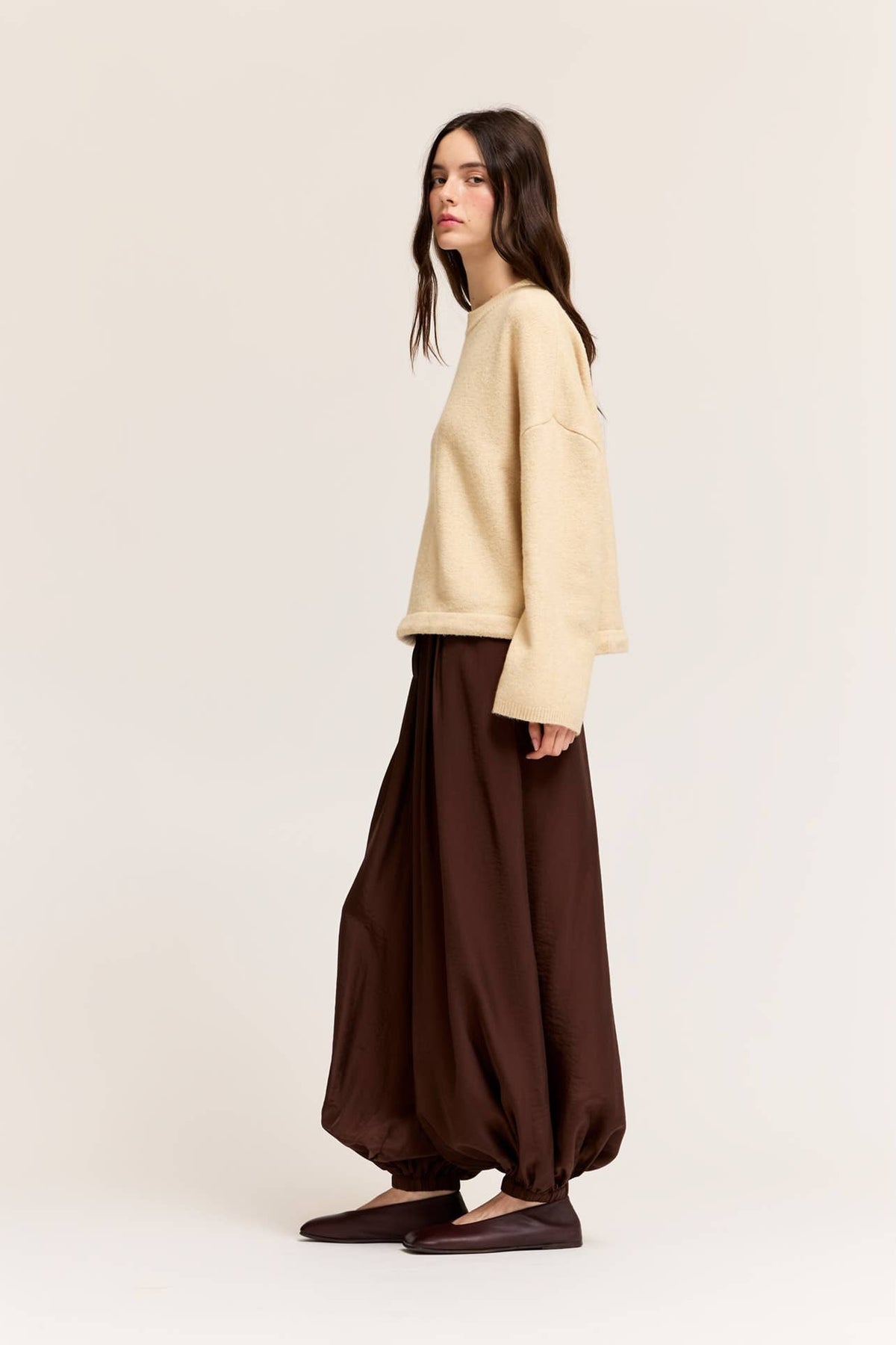 A woman modeling a cream-colored oversized long-sleeve top and deep bark satin harem pants against a plain beige studio background.
