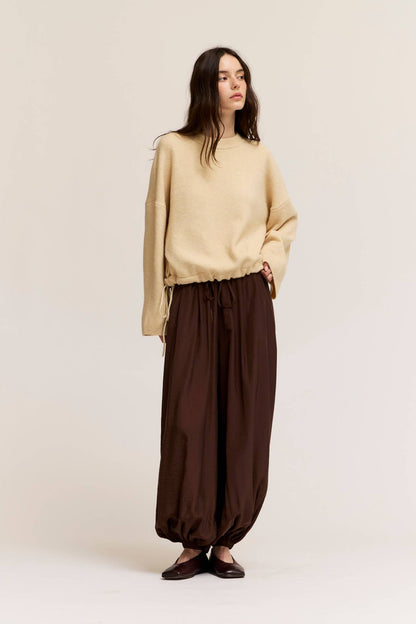 A woman stands against a plain light beige studio background wearing an oversized cream knit sweater and loose deep bark satin harem pants with dark flat shoes.