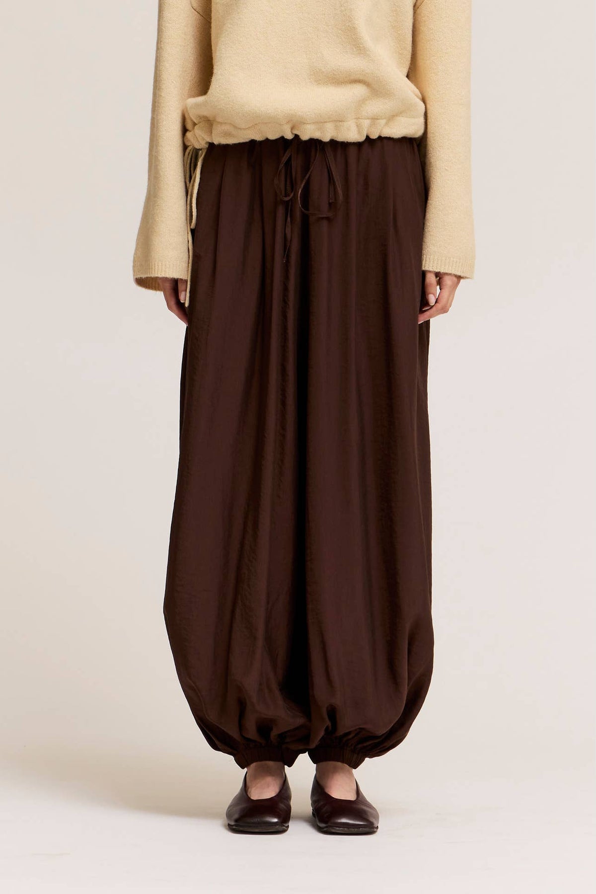 A person wearing deep bark satin harem pants with gathered cuffs, paired with a cream sweater and dark flats, shown against a plain light beige studio background.