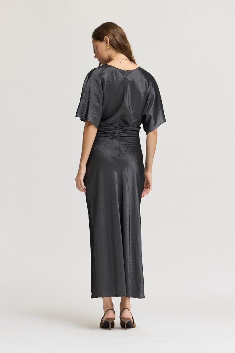 A woman viewed from the back wearing a dark granite wash gray satin midi dress with short flutter sleeves, a gathered waist, and a draped open back, paired with black heeled sandals against a light gray studio background.