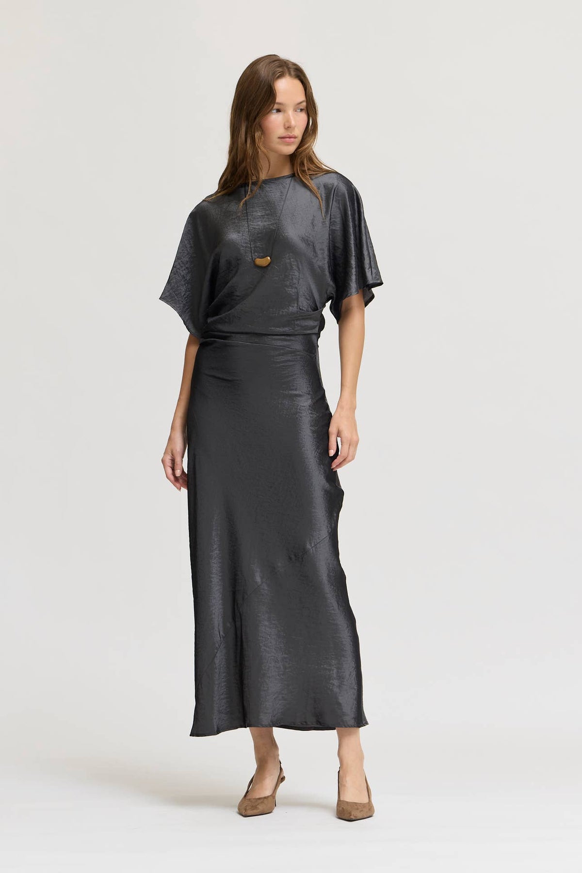 A woman modeling a dark granite wash gray, short-sleeved wrap-style midi dress with a metallic sheen, paired with brown pointed-toe heels, against a light gray studio background.