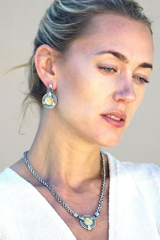 A woman wearing a matching silver-toned necklace and earring with a gold and silver circular coin pendant, shown in a close-up portrait against a plain beige background.