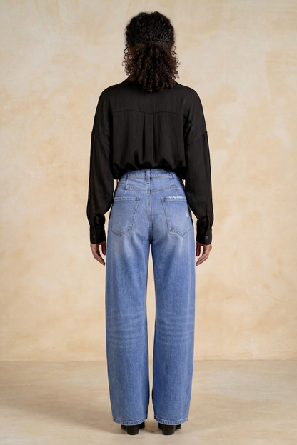 A model viewed from the back wearing a black long-sleeve blouse and high rise barrel denim jeans, standing against a textured beige studio backdrop.