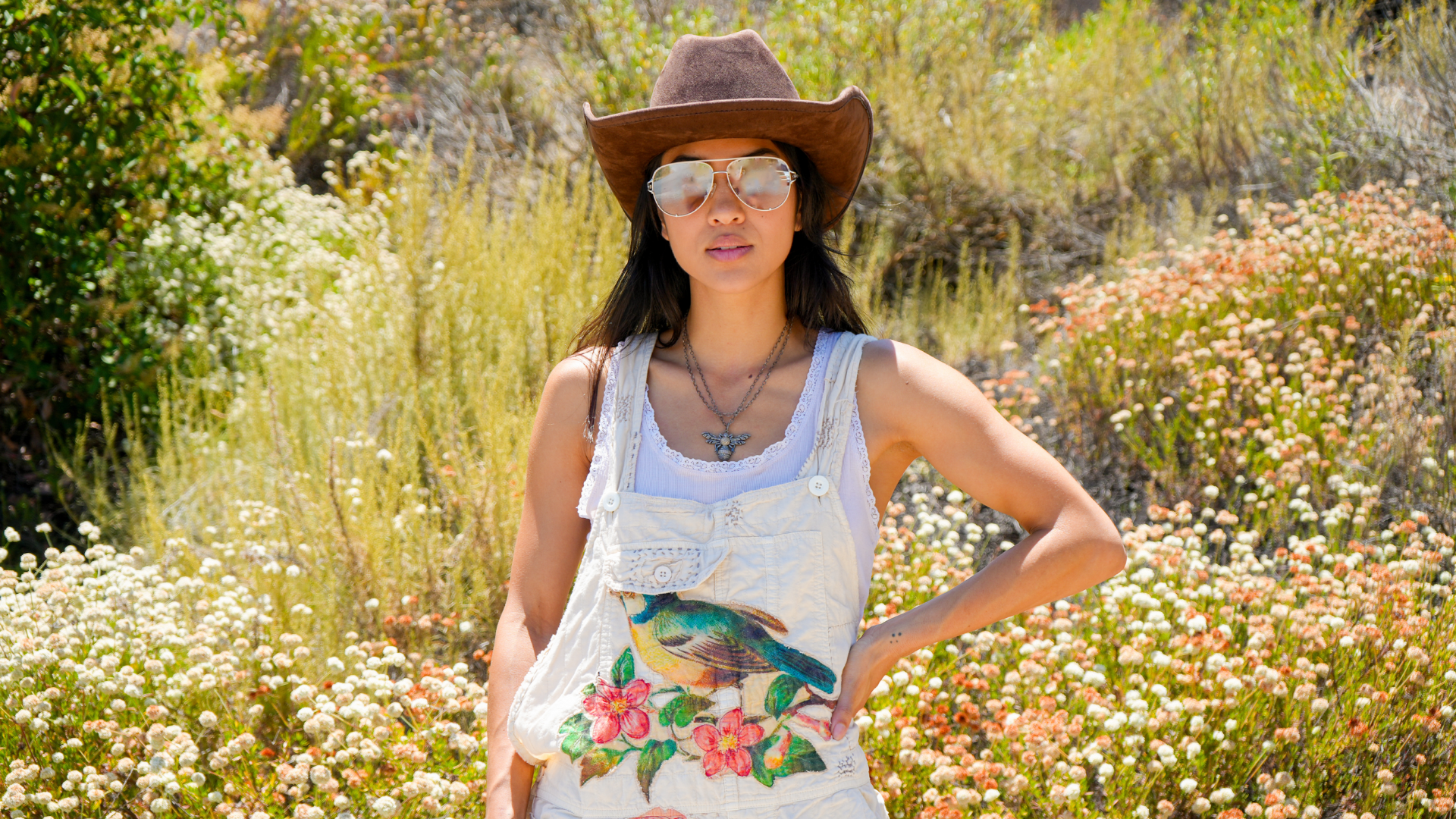 Woman wearing a cowboy hat and floral overalls standing in a natural setting with flowers and greenery.