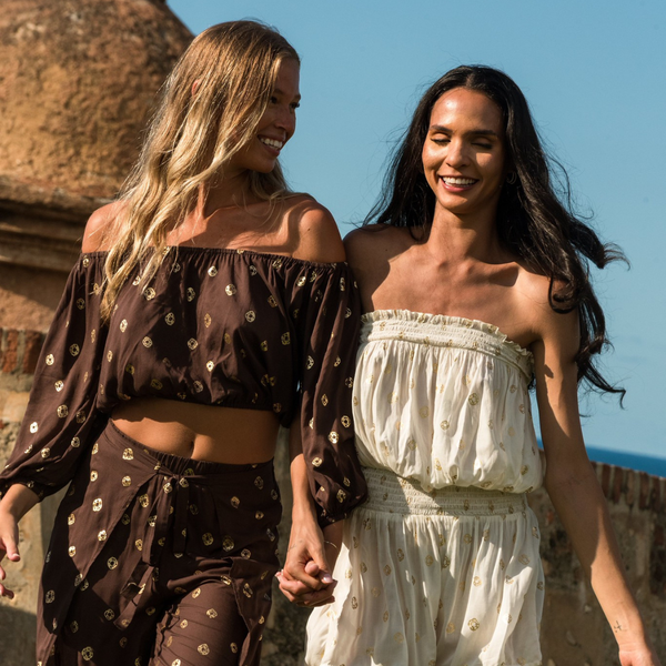 Two women in patterned outfits smile and walk together outdoors near a stone building under a clear blue sky.