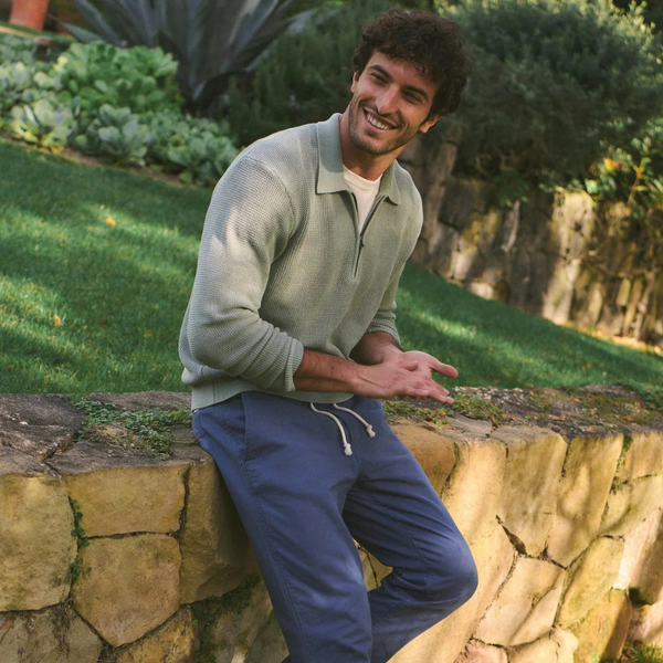 Smiling man sits on a stone wall in a garden, wearing a green sweater and blue pants surrounded by plants.