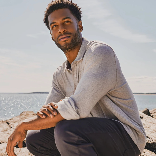 A man crouching outdoors on rocks near the water, wearing a light gray long-sleeve button-up shirt and dark pants.