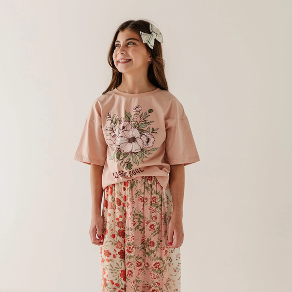 A young girl wearing a dusty pink short-sleeve t-shirt with a large floral graphic on the front and matching floral pants, standing against a plain off-white background.