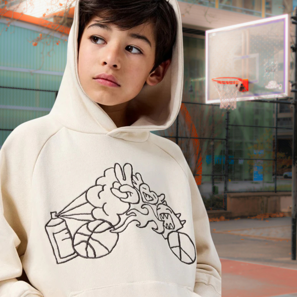 Young person in a white hoodie stands in a basketball court with a hoop visible in the background.
