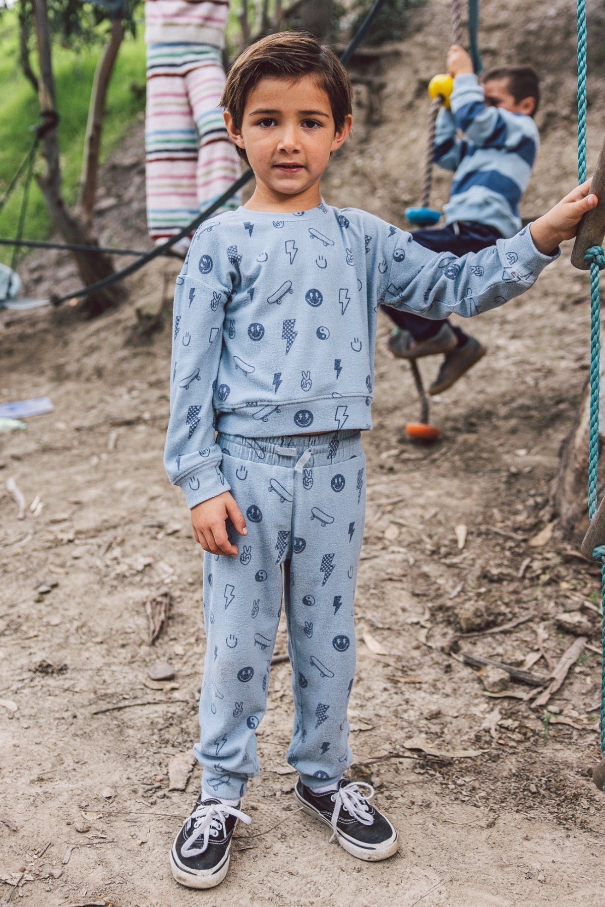 A young boy wearing a light blue lounge set with a dark blue cartoon lightning-bolt pattern stands in a dirt playground, holding onto a wooden climbing frame. Another child is visible in the background on a swing.