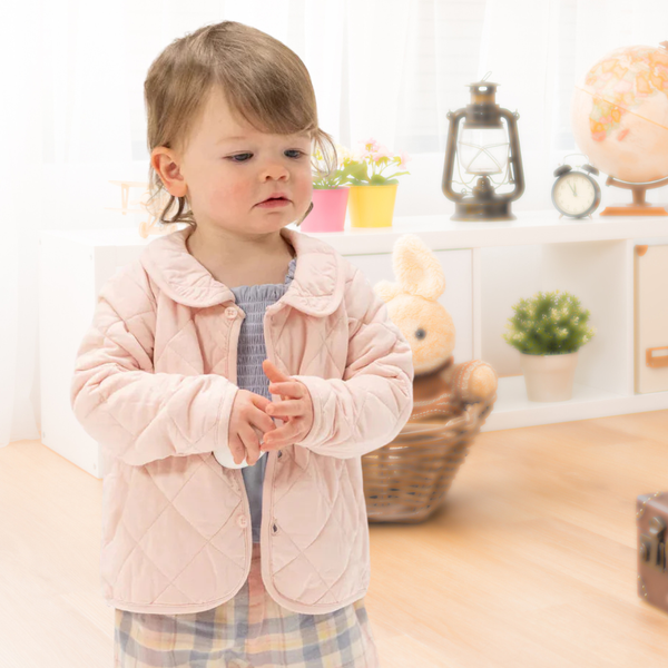 Young child in a pink jacket stands indoors near a shelf with a toy bunny, plants, lantern, clock, and globe.