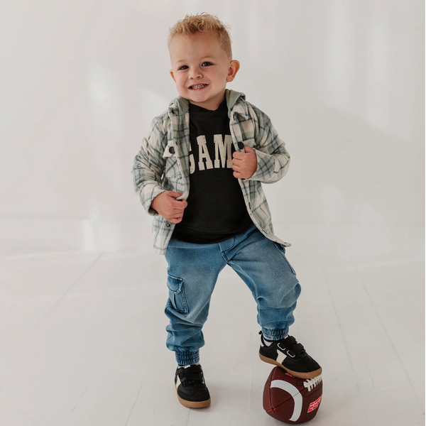 A toddler boy wearing a black graphic t-shirt, a gray and white plaid flannel hoodie, blue jeans, and black sneakers, standing on a white football in a studio setting.