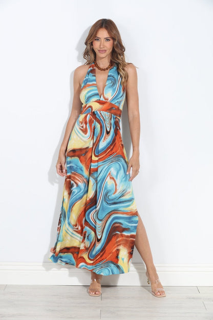 A woman modeling a sleeveless halter-neck maxi dress with a vibrant blue, orange, and yellow marbled sahara pattern, standing against a white wall.