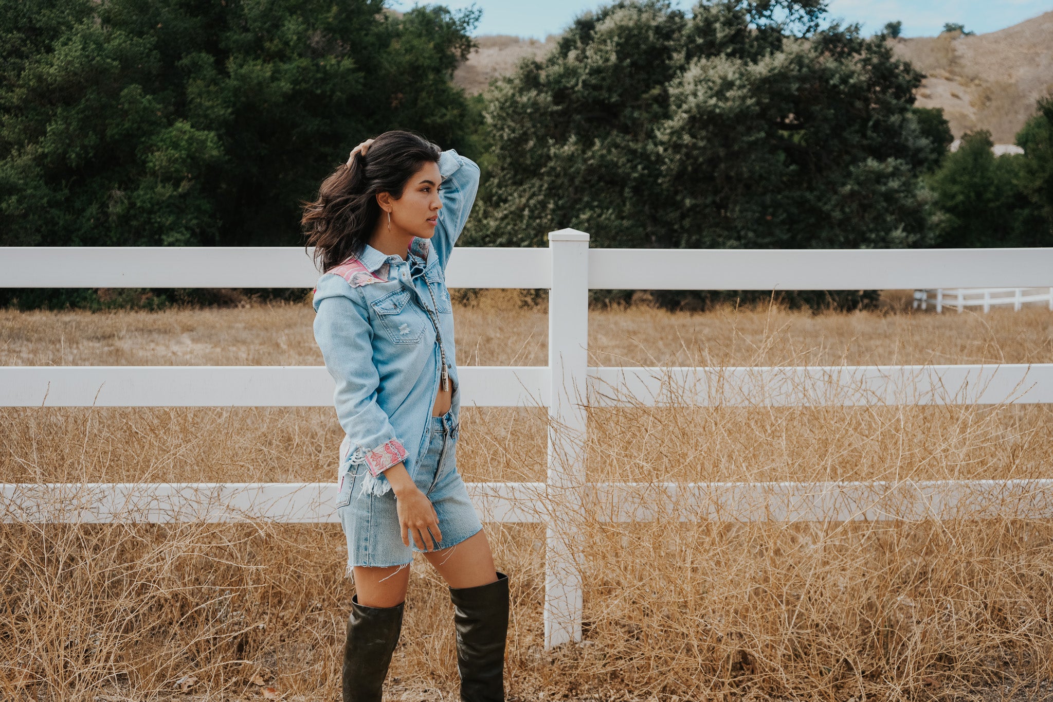Woman in a denim outfit and knee-high boots standing in a field with a white fence and trees in the background.