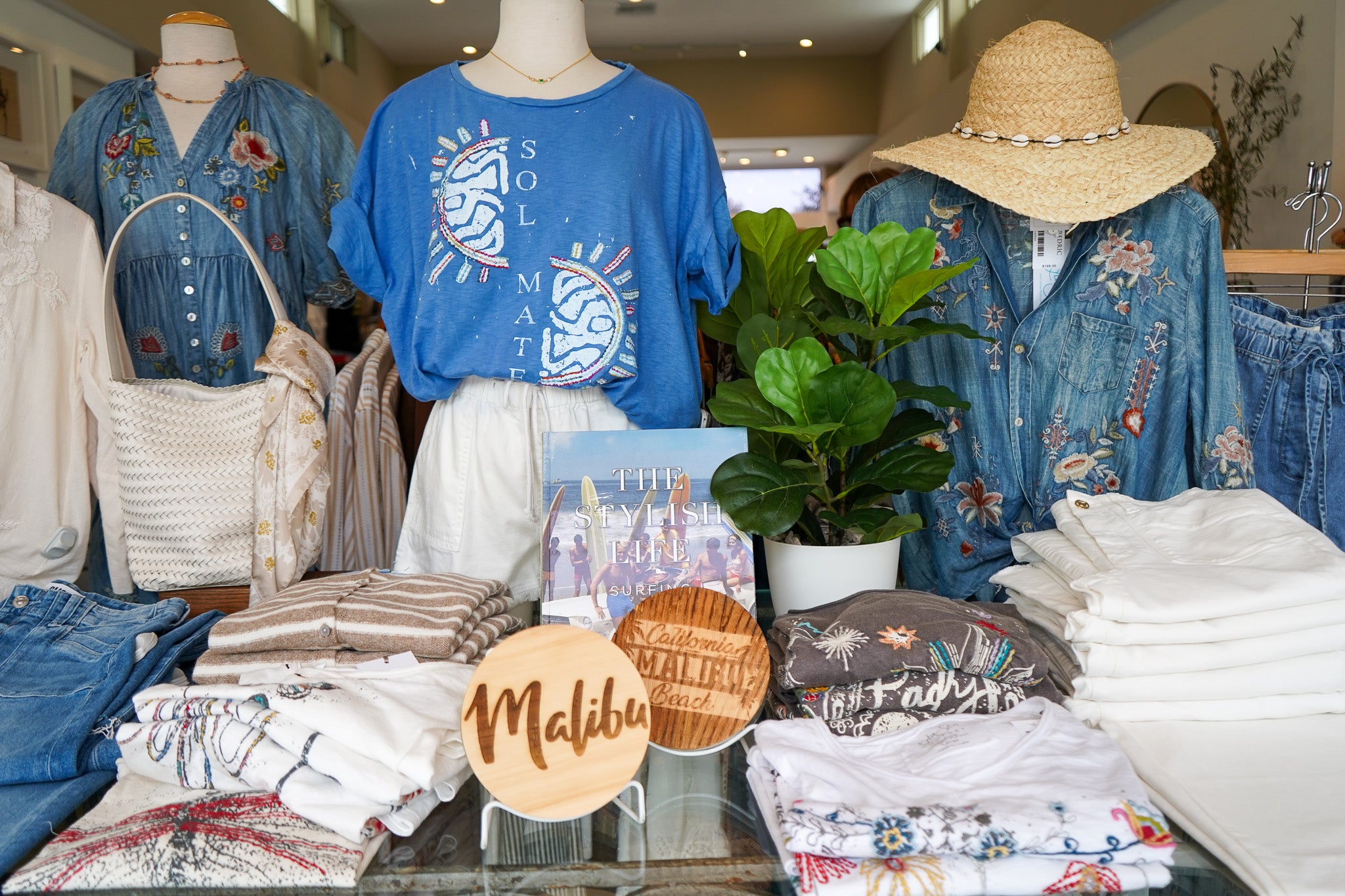 A boutique display featuring a blue t-shirt with a white sun graphic alongside other clothing items, folded white denim and shirts, a potted green plant, and wooden Malibu-themed signs on a glass table.