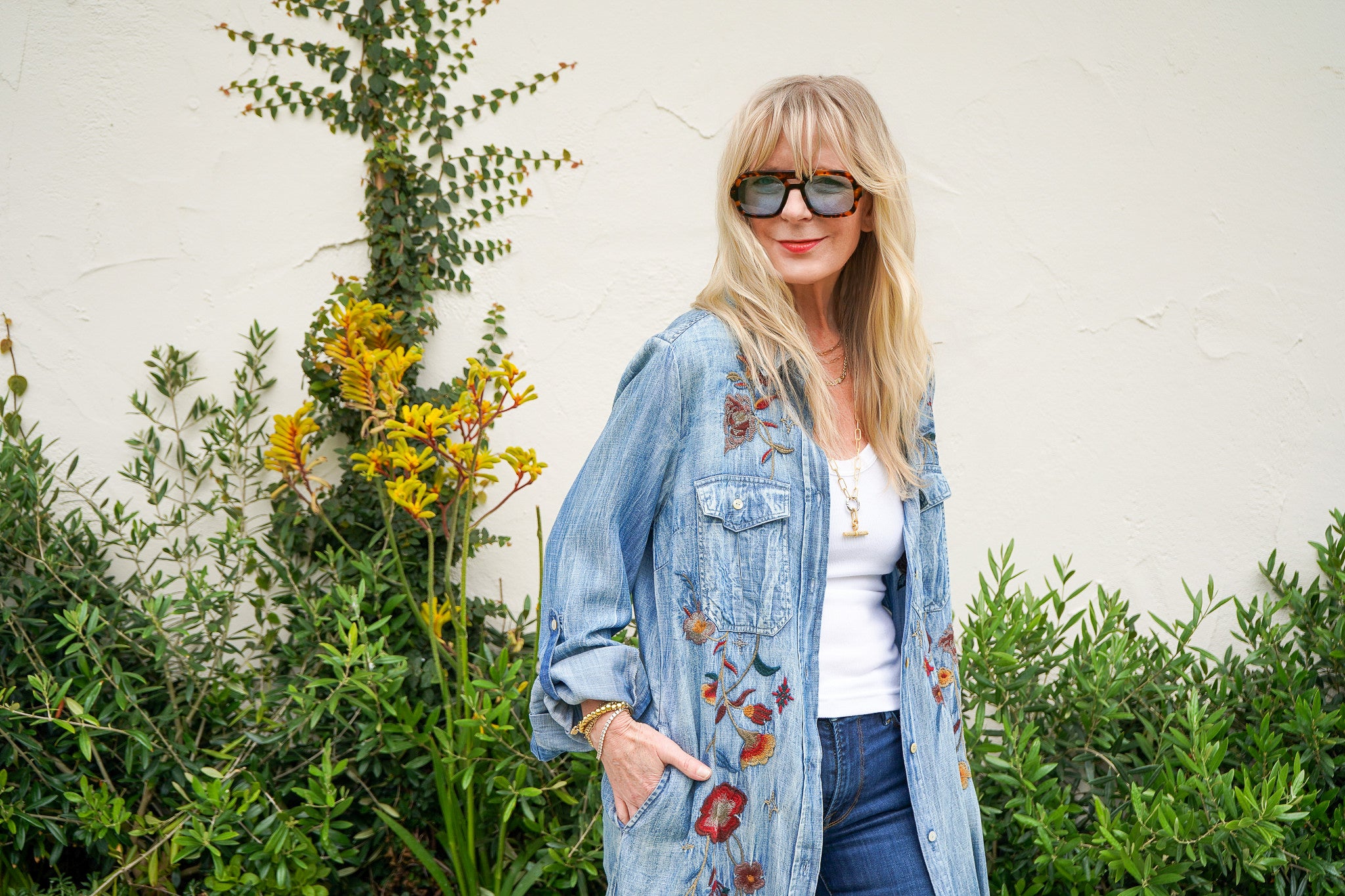 A woman with blonde hair and sunglasses wearing a light blue denim jacket with red and orange floral embroidery, posing outdoors against a white wall and green plants.