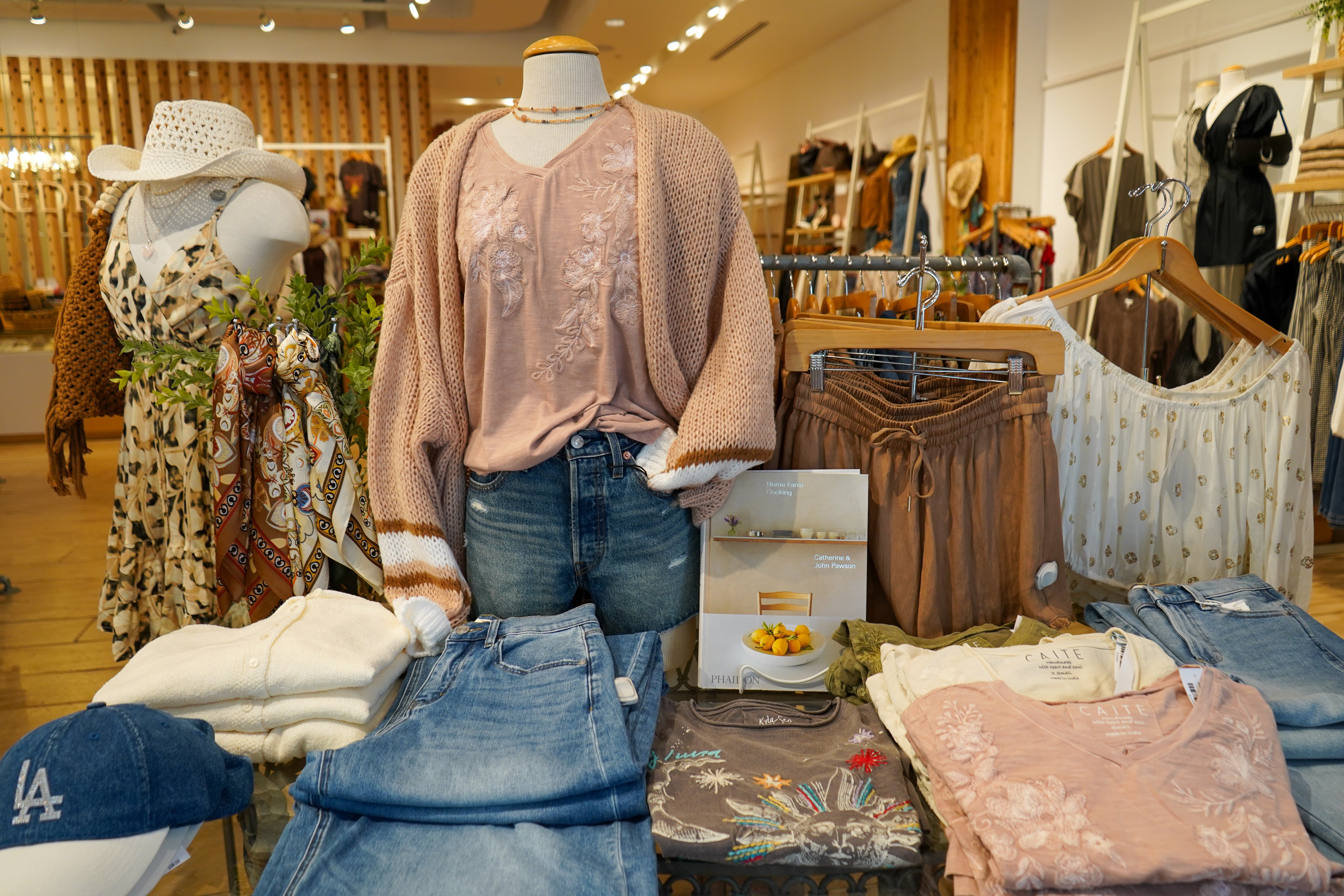A retail clothing display inside a boutique featuring a mannequin dressed in a dusty pink knit cardigan over a matching embroidered top and blue jeans. Folded garments, hangers, and other apparel are arranged around the display table in a well-lit store interior.