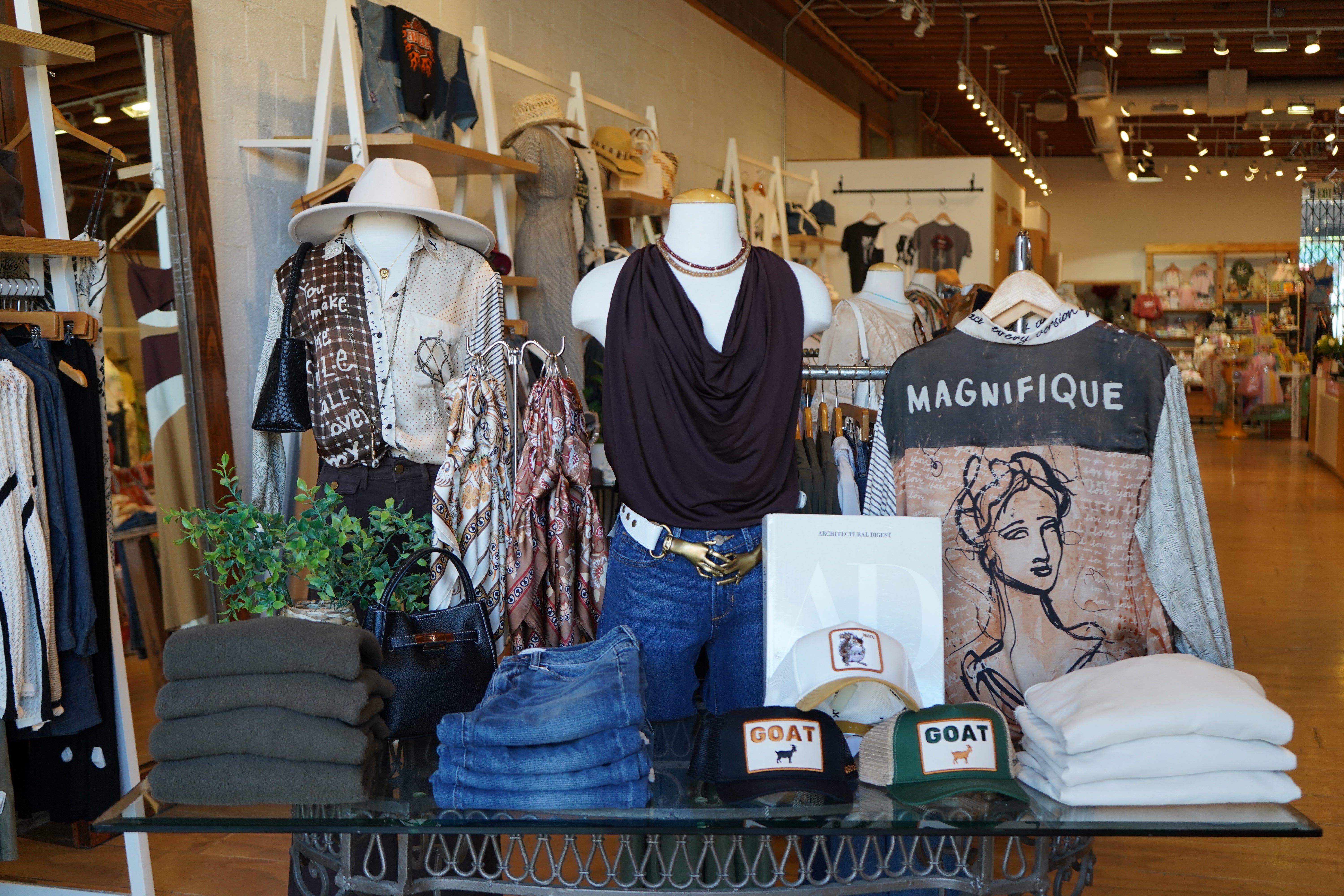 A retail clothing display inside a store, featuring mannequins and racks with shirts, jeans, folded garments, and a sweatshirt printed with 'MAGNIFIQUE' and a woman's face. A glass table in the foreground holds folded clothes and three caps, including one that reads 'GOAT'.