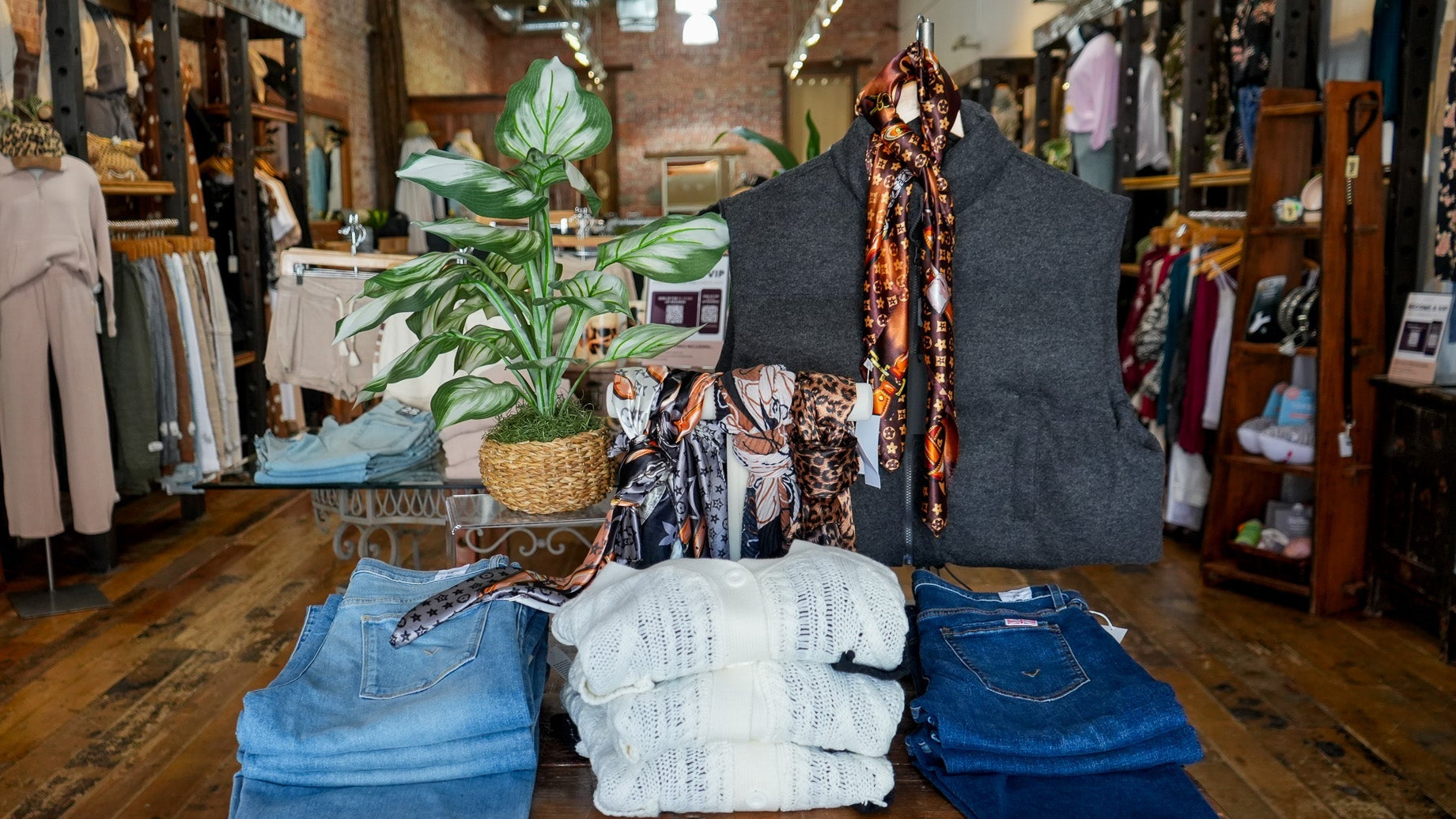 Display of jeans and a vest on a table in a clothing store with plants and shelves in the background.