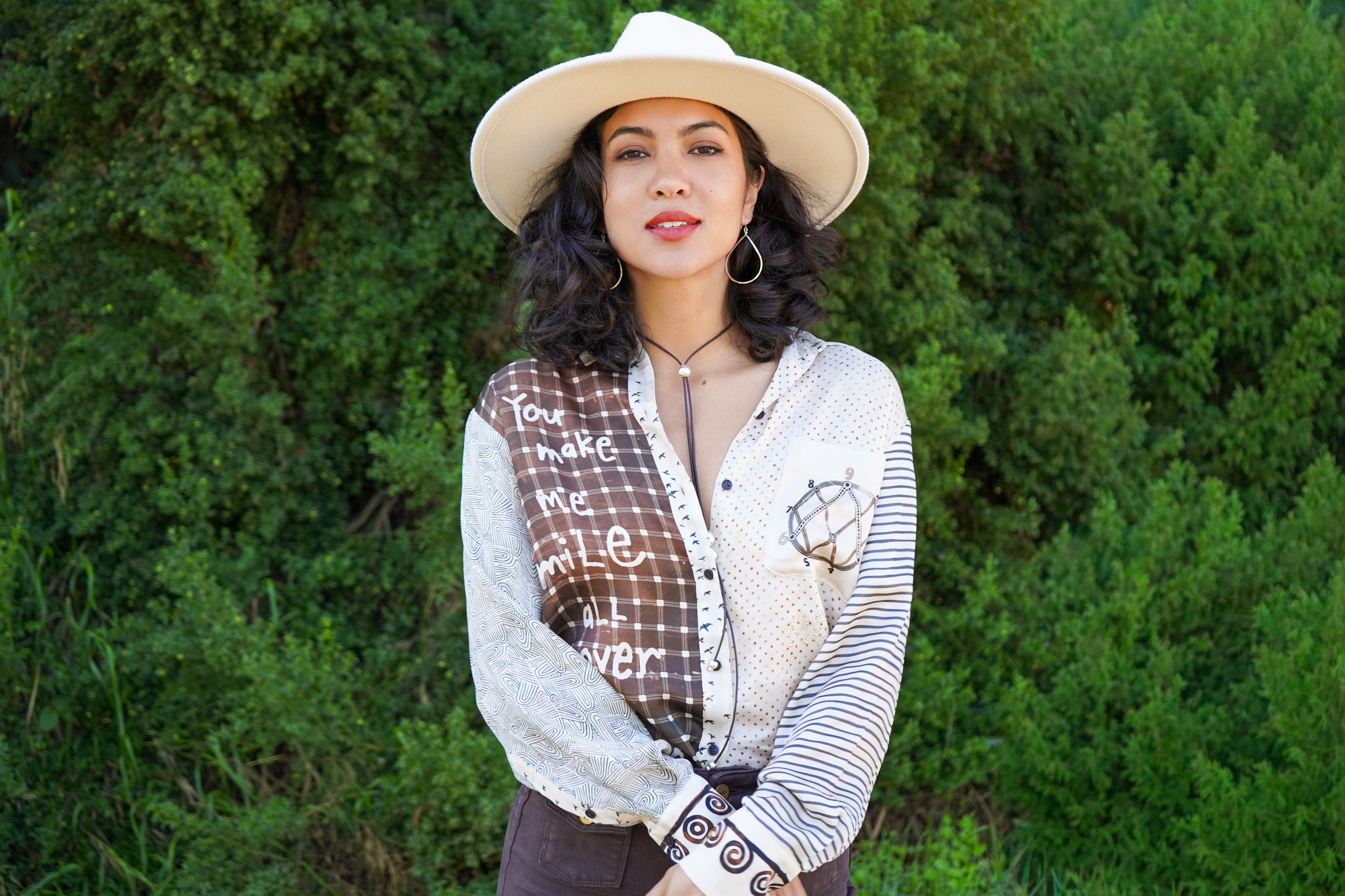 A woman wearing a cream wide-brim hat, a patchwork button-up shirt with brown plaid and striped sections, and a brown vest, standing outdoors against a background of green bushes.