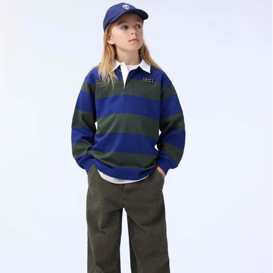 A boy wears a striped polo, blue hat and dark jeans on a white background