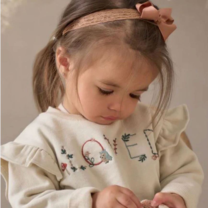 Child wearing a beige sweater with embroidered designs and a pink headband.