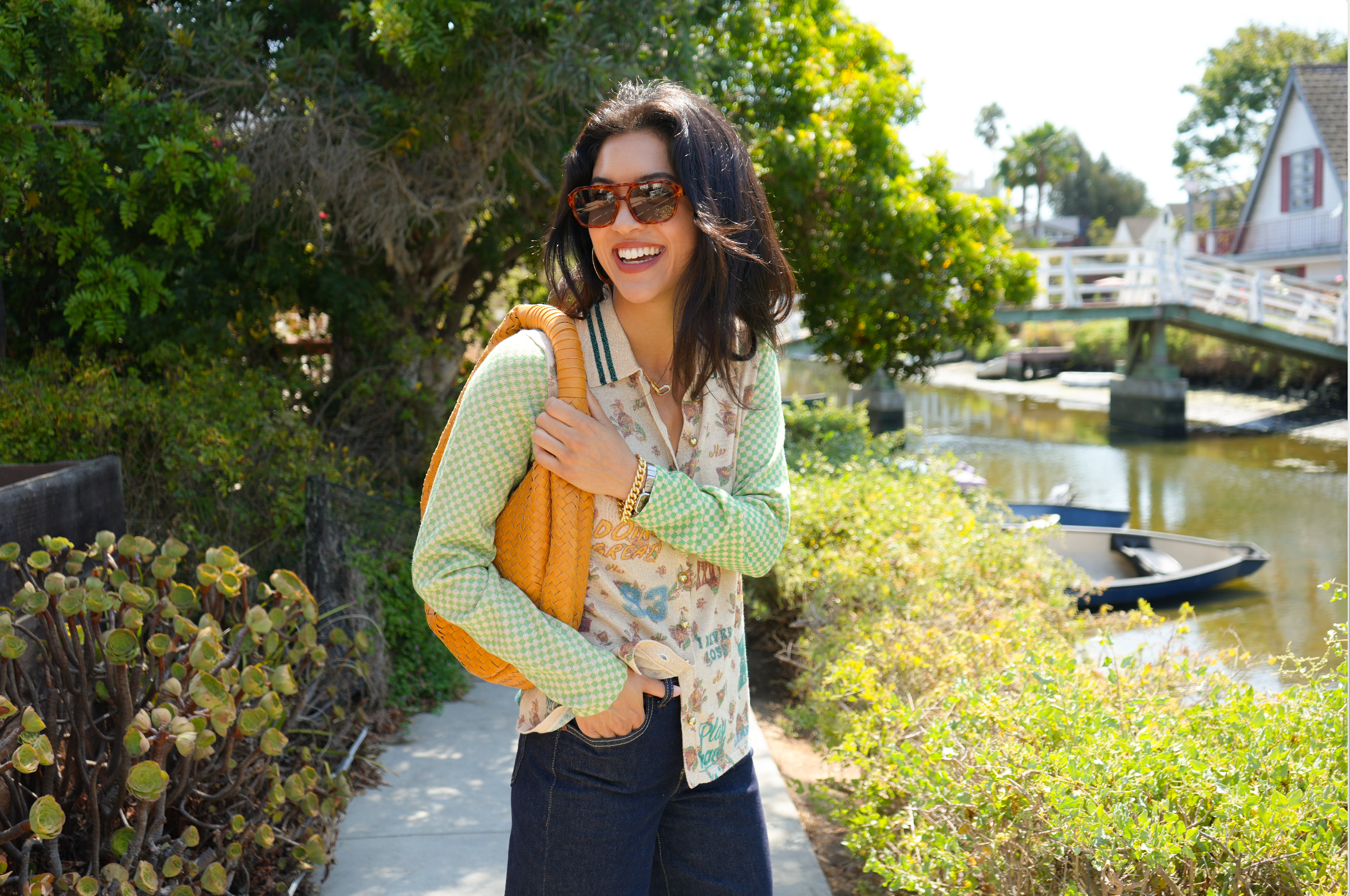 A woman holding a yellow handbag and wearing sunglasses, a sweater and jeans stands on a canal walk way