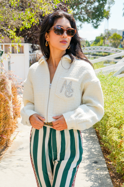 Woman wearing a cream jacket with a logo and green and white striped pants outdoors.