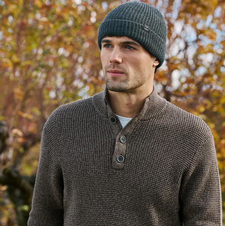 Man wearing a brown knitted sweater and gray beanie outdoors with blurred autumn trees in the background