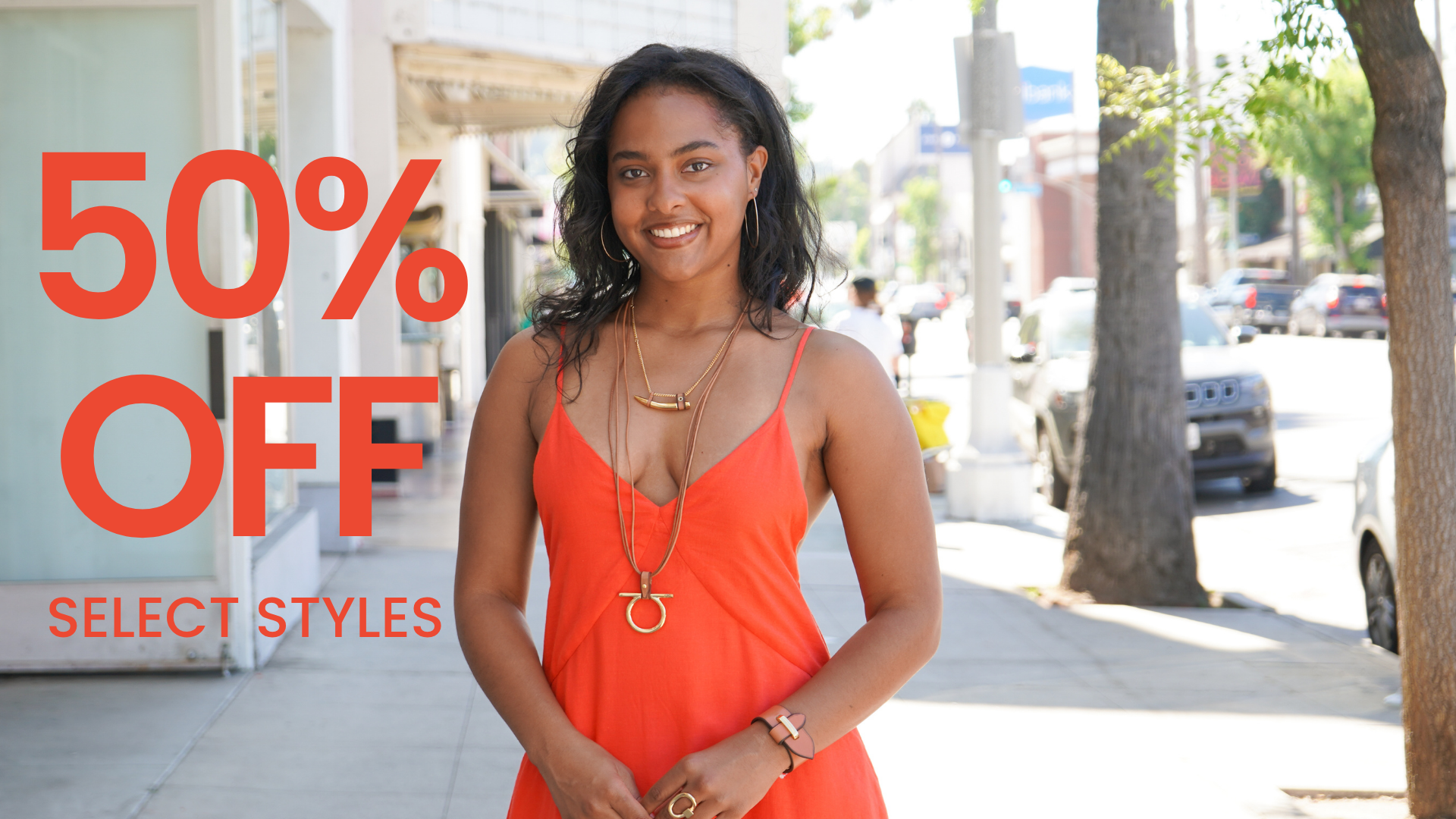 A woman wearing a bright orange sleeveless dress stands on a city sidewalk with large red text overlay reading '50% OFF' and smaller text 'SELECT STYLES'.