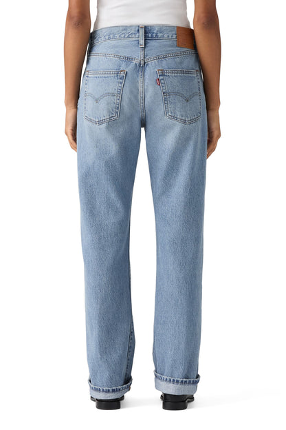 Rear view of a person wearing light-wash hallowed hour selvedge blue denim jeans with a white top and black shoes, shown against a plain white background.