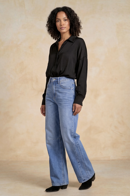 A model wearing a black long-sleeve button-up shirt and high rise barrel jeans, standing against a textured beige studio backdrop.