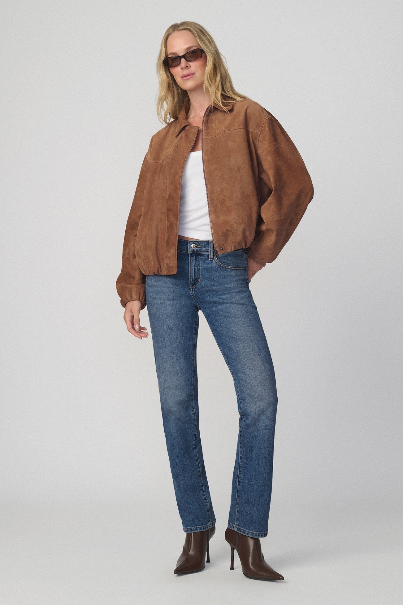 A woman modeling a brown suede-like cropped jacket over a white top, paired with amalfi colored blue jeans and brown heeled ankle boots, against a light gray studio background.