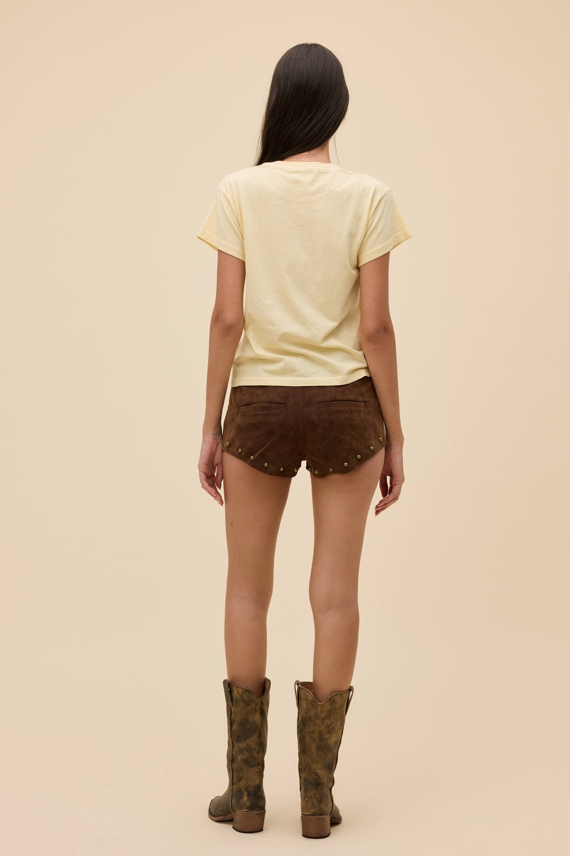 Back view of person wearing a sheer yellow top and brown shorts with studded details on a beige background