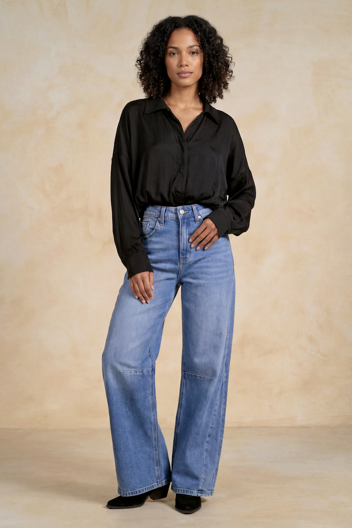 A woman modeling a black long-sleeve button-up shirt and high-waisted, barrel light blue jeans against a textured beige studio backdrop.