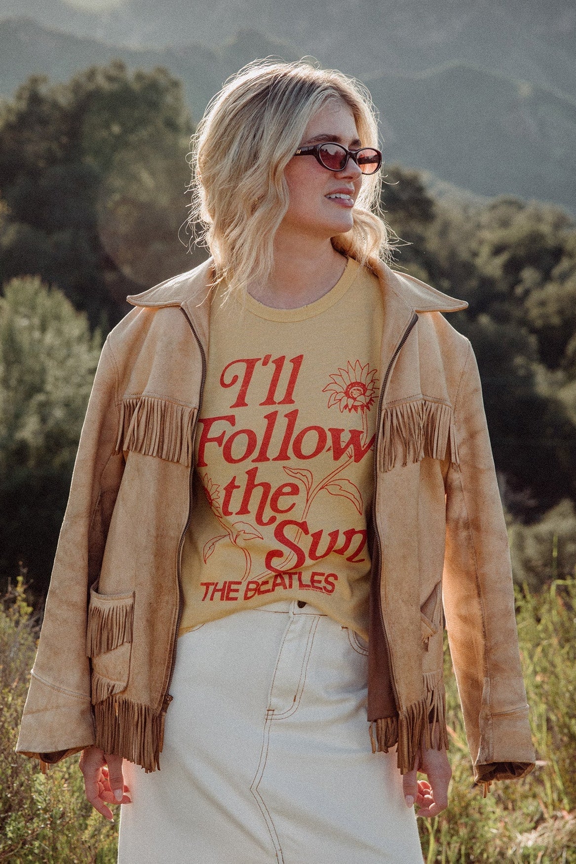 Woman wearing a tan fringed jacket over a dusty citron Beatles t-shirt in a natural setting
