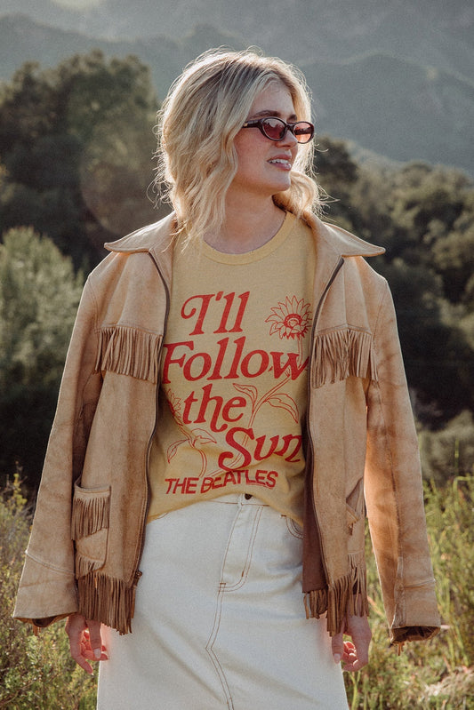 Woman wearing a tan fringed jacket over a dusty citron Beatles t-shirt in a natural setting